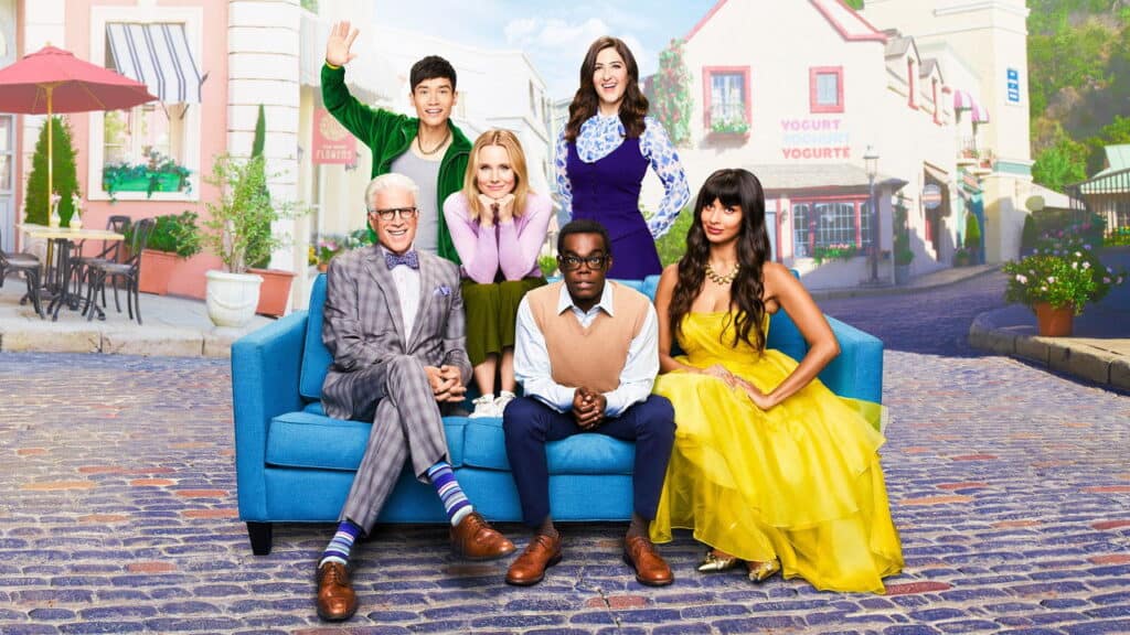 Ensemble cast seated together in a bright, whimsical town setting, reflecting the thoughtful humor and heart of philosophical comedy shows like The Good Place.