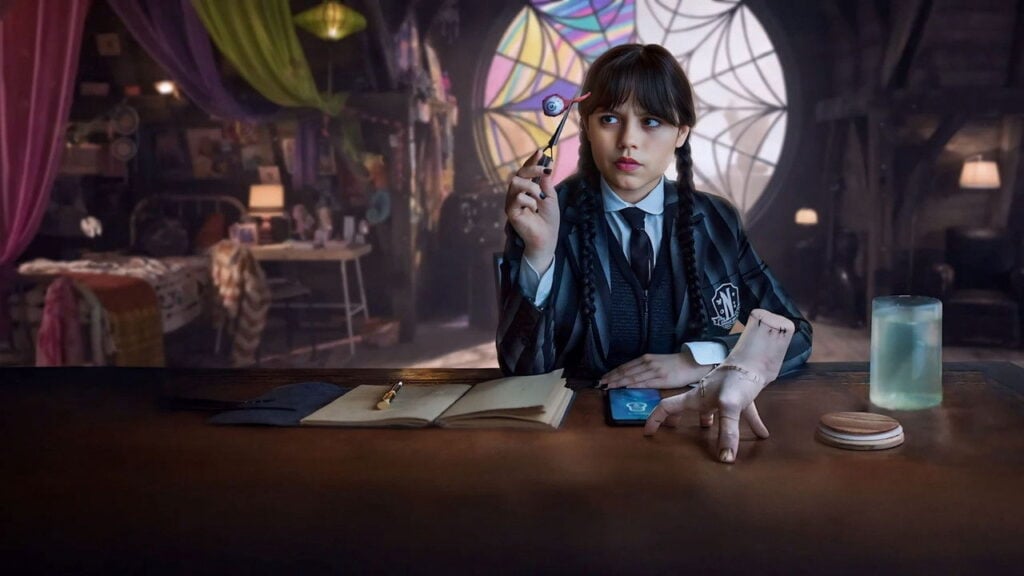 A teenage girl in a dark school uniform sits at a desk inside a dimly lit room, holding a strange object while studying it with quiet focus