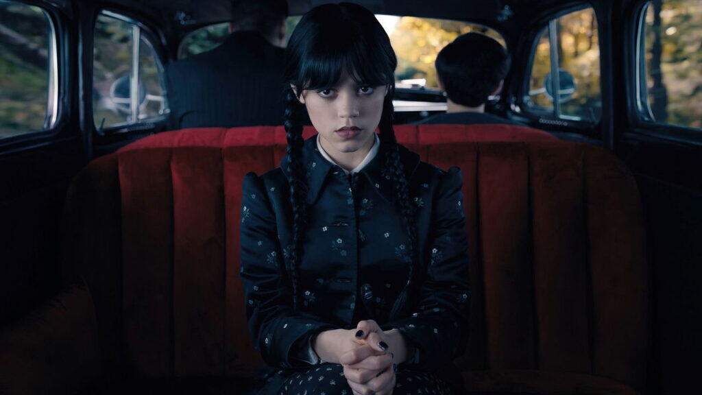A pale teenage girl with braided hair sits upright in the back seat of a vintage car, staring forward with a calm, unreadable expression