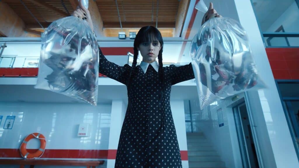 A darkly dressed teenage girl with braided hair holds two clear bags containing live piranhas inside a modern indoor pool facility