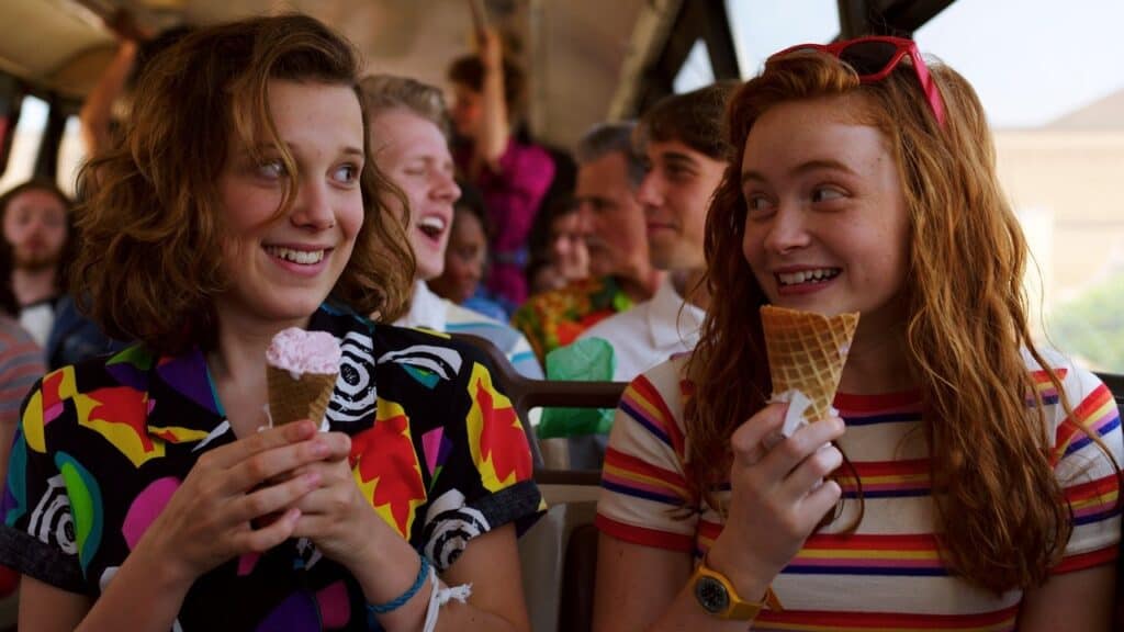 Two friends smile at each other while holding ice cream cones on a crowded bus, sharing a lighthearted moment in Stranger Things.