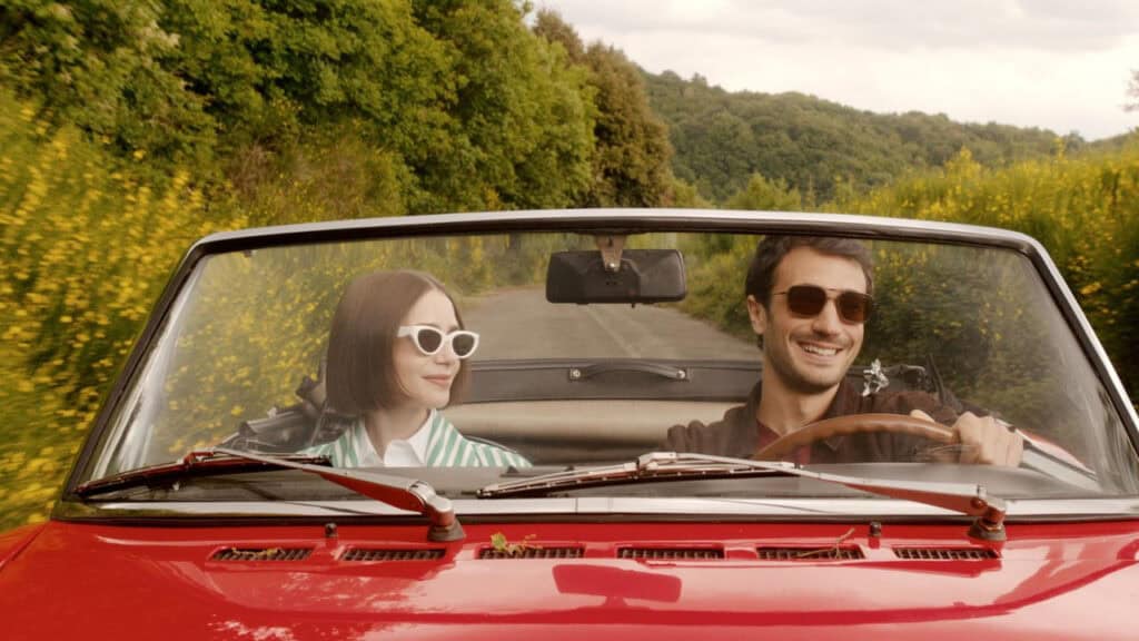 A couple driving a red convertible through the countryside, smiling and enjoying the ride.