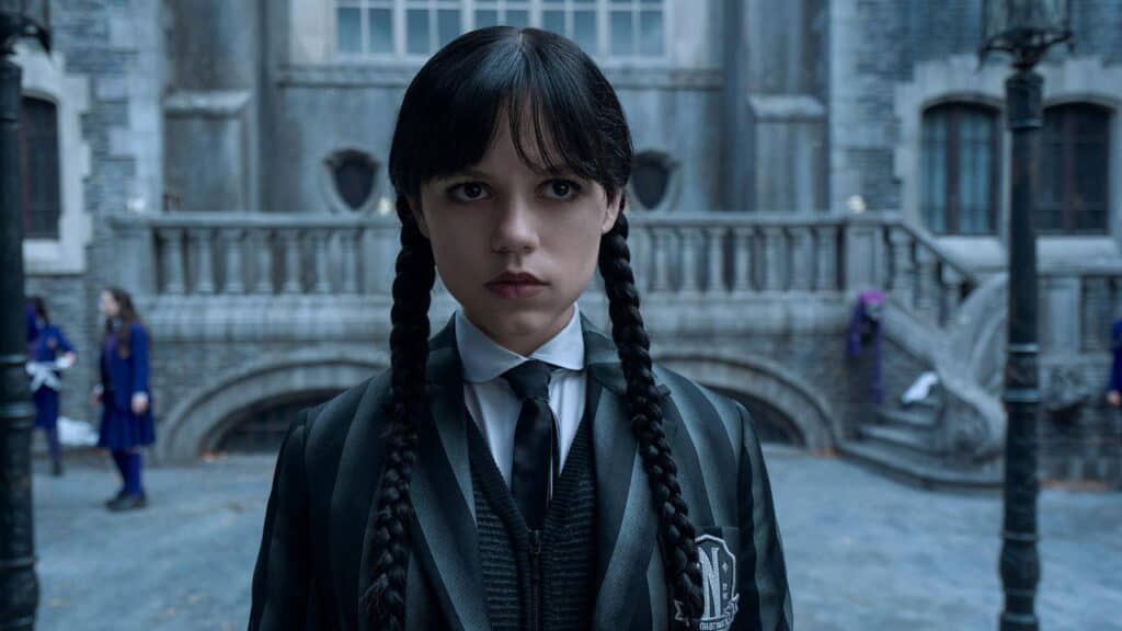 A teenage girl in a black school uniform stands motionless in a stone courtyard of a gothic academy, looking directly ahead