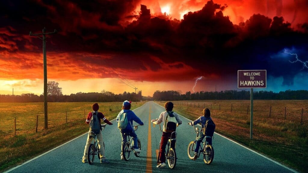 Four kids on bicycles stop in the middle of a rural road beneath a dramatic sky near a sign welcoming visitors to Hawkins in Stranger Things.