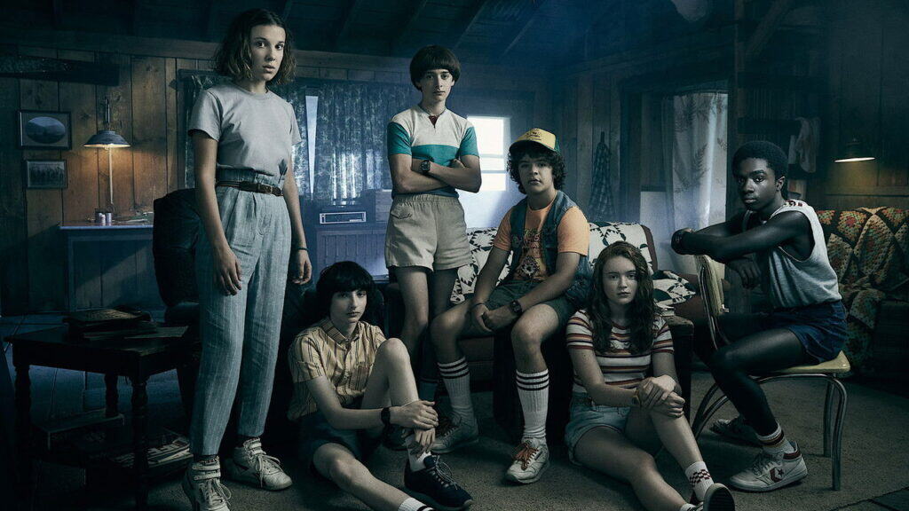 A group of young friends stand together inside a dimly lit cabin, tense and uncertain as danger closes in during Stranger Things.