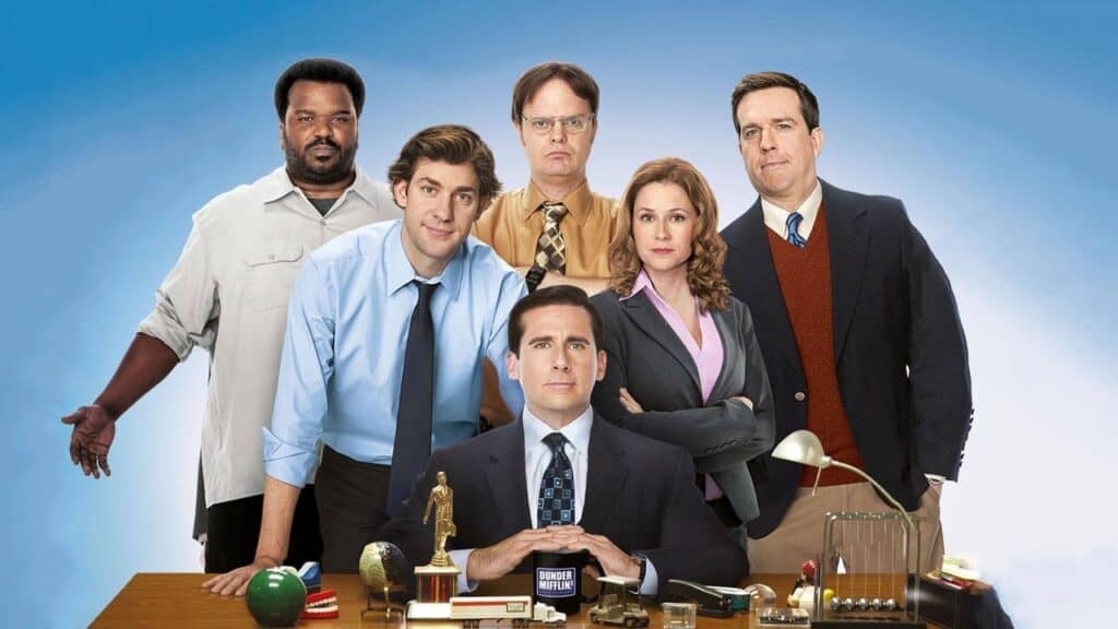 Group portrait of The Office cast standing behind an office desk with props and paperwork in a studio setting