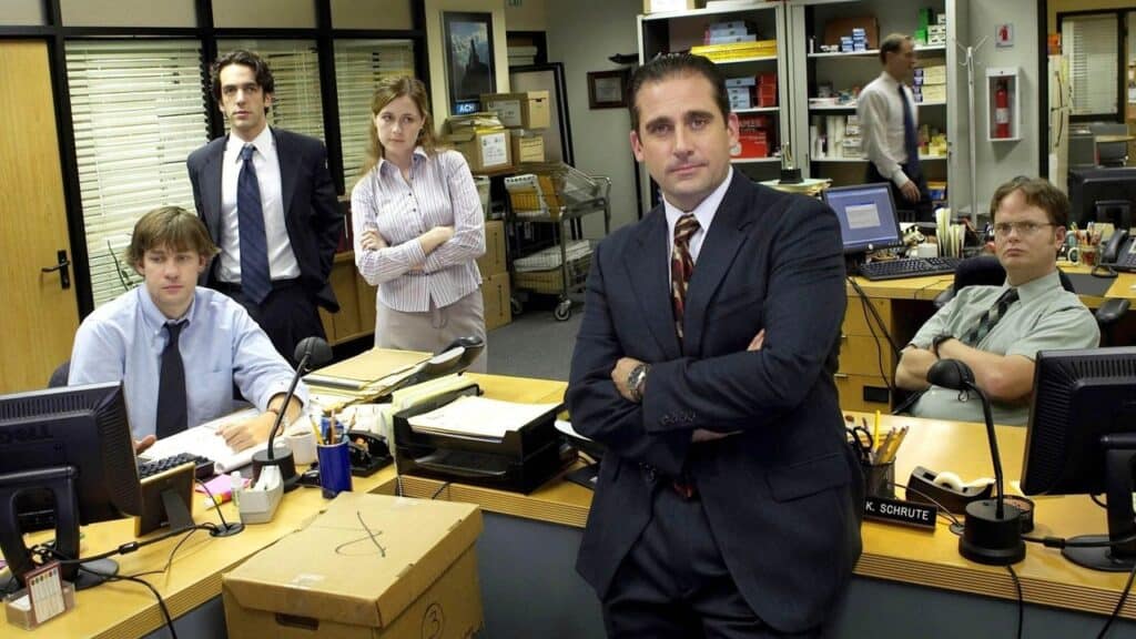 Office staff gathered in a conference room with desks, paperwork, and computers during a tense workplace moment