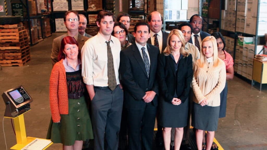 Full cast of The Office standing together inside a warehouse with shelves and pallets in the background