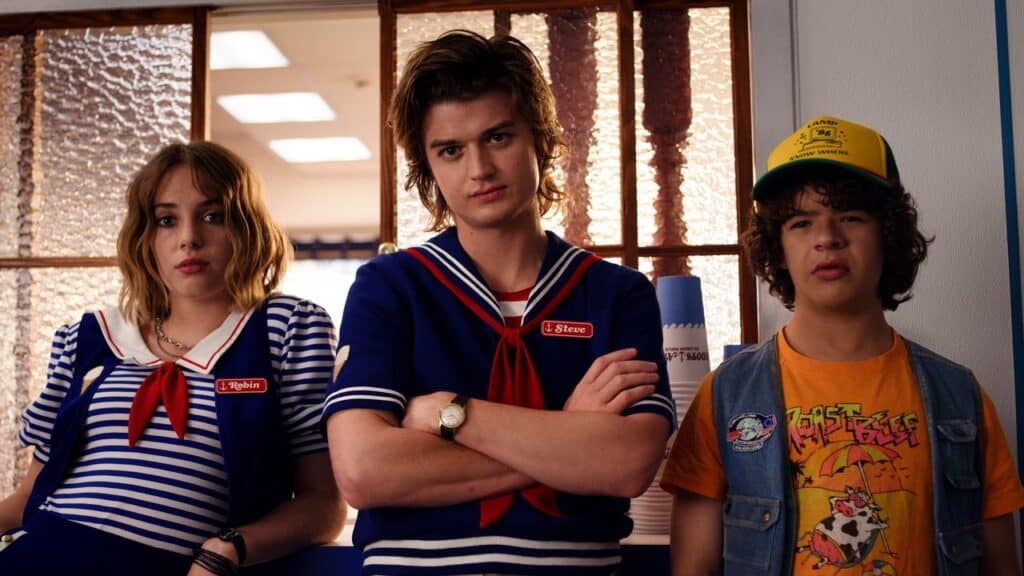 Three teens stand inside an ice cream shop wearing uniforms, their expressions serious despite the bright setting in Stranger Things.