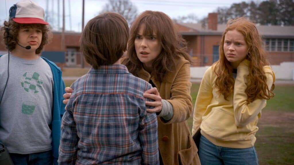 A worried mother grips her young son’s shoulders on a school field while two other children look on, capturing fear and confusion as supernatural events begin to disrupt everyday life.