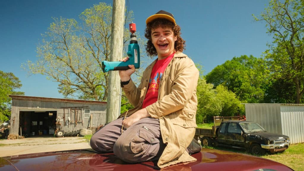 A teenage boy smiles while holding a power drill outdoors in a sunny rural setting in Stranger Things.