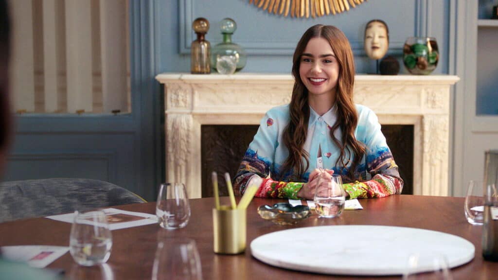 Emily sits at a modern dining table in a blue room smiling during a professional dinner meeting surrounded by glassware and decor