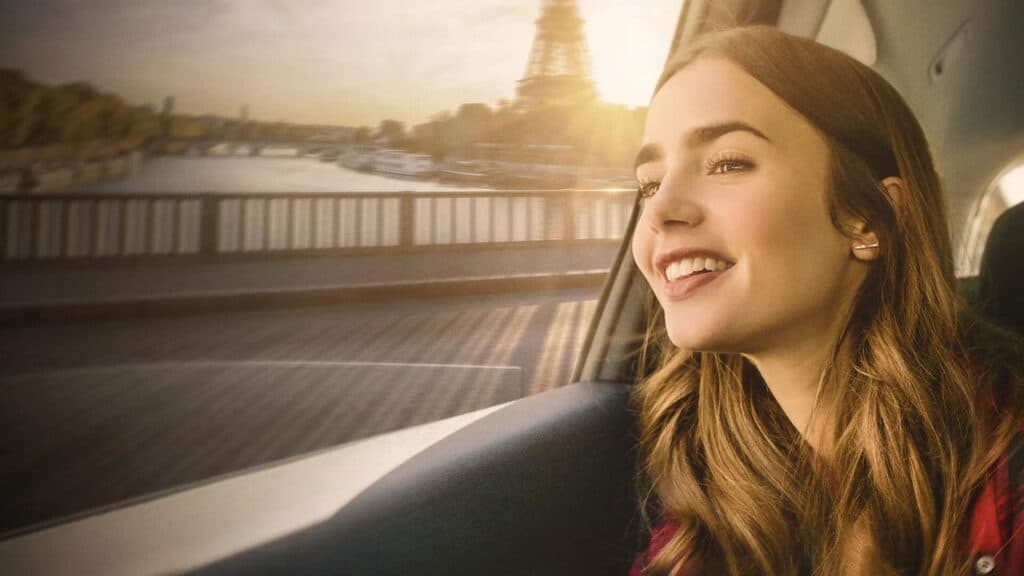 Emily smiles out the car window while riding across a bridge in Paris with the Eiffel Tower glowing in the background at sunset