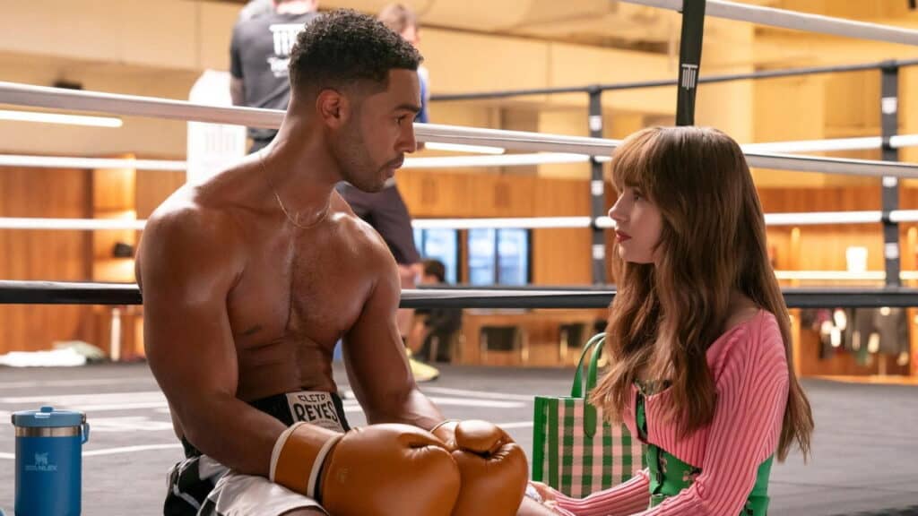 Emily sits ringside at a boxing gym in Paris talking to a shirtless boxer wearing gloves during a quiet training moment
