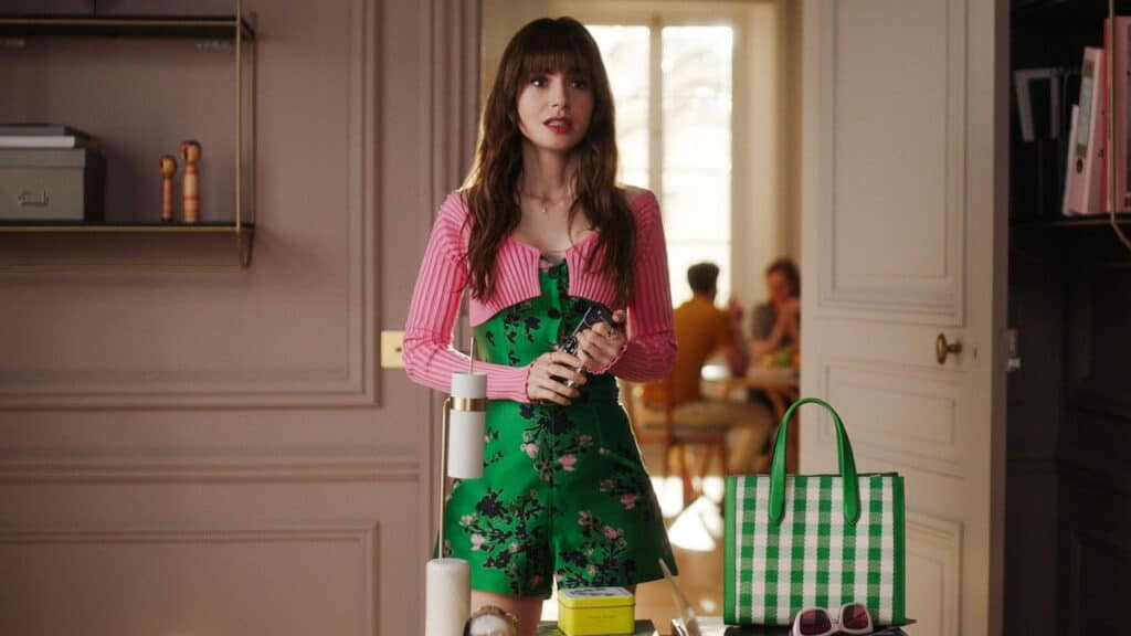Emily stands in her Paris apartment wearing a green floral romper and pink cardigan while holding her phone, surrounded by stylish decor and accessories