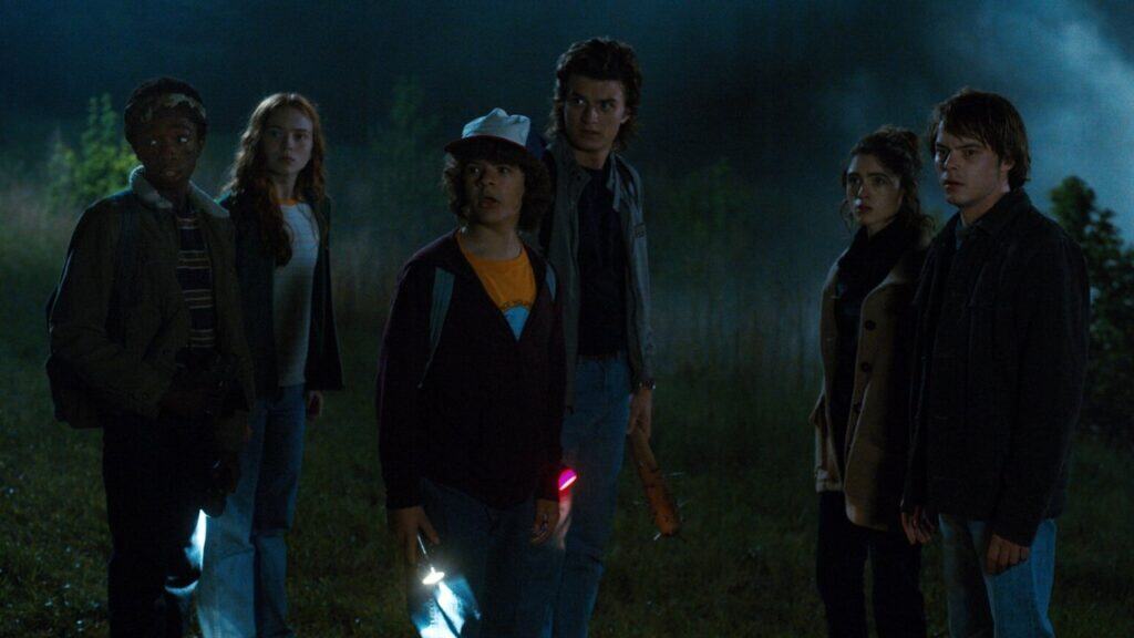 A group of teenagers stand in a dark field at night holding flashlights and weapons, united in fear and determination as they prepare to confront a hidden supernatural danger.