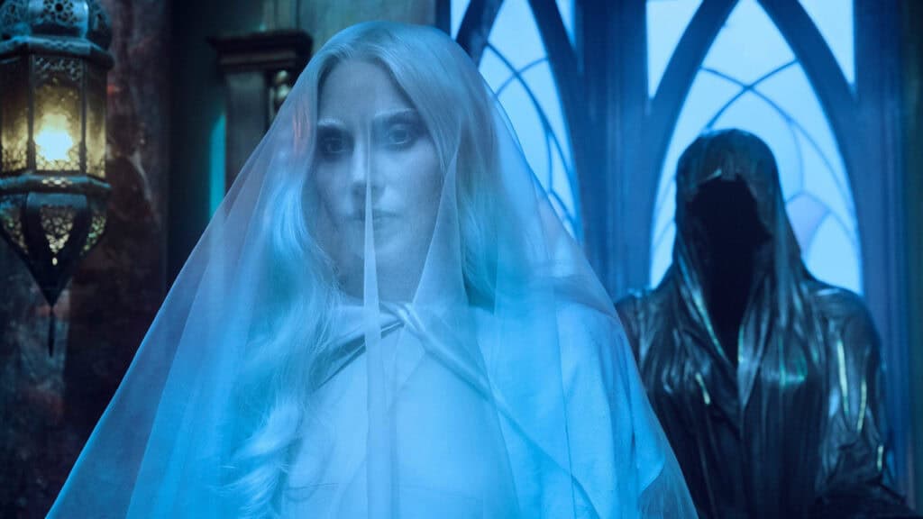 A ghostly woman in a veil stands in a dim blue hallway inside Nevermore Academy, creating a mysterious and eerie atmosphere in Wednesday.