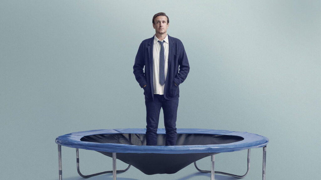 A man wearing a loose jacket and tie stands still on a trampoline in a pale blue room, looking upward with a thoughtful expression.