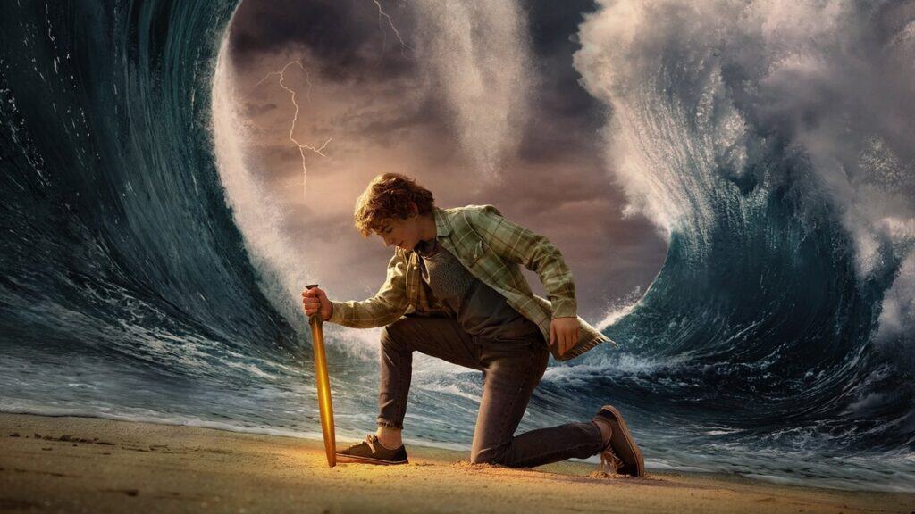 Percy Jackson kneels in the sand holding a glowing sword as massive ocean waves rise and part behind him