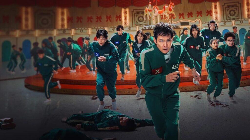 A group of contestants in green tracksuits run in panic during a deadly children’s game, with fallen players on the floor and fear visible across their faces.