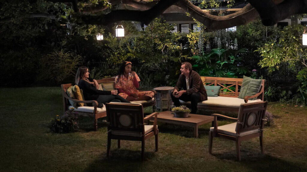 A small group of adults sit together in a softly lit backyard at night, talking casually on outdoor furniture beneath trees and lanterns.