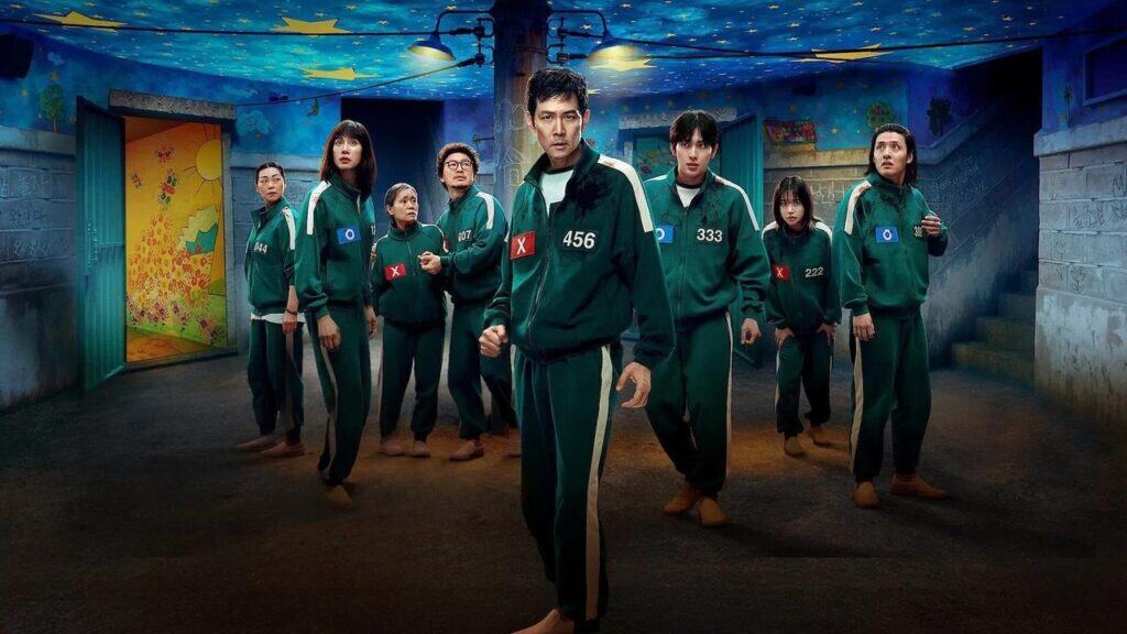 Several main contestants stand in a dimly lit room with painted walls, looking tense and uncertain as they prepare for another dangerous challenge.