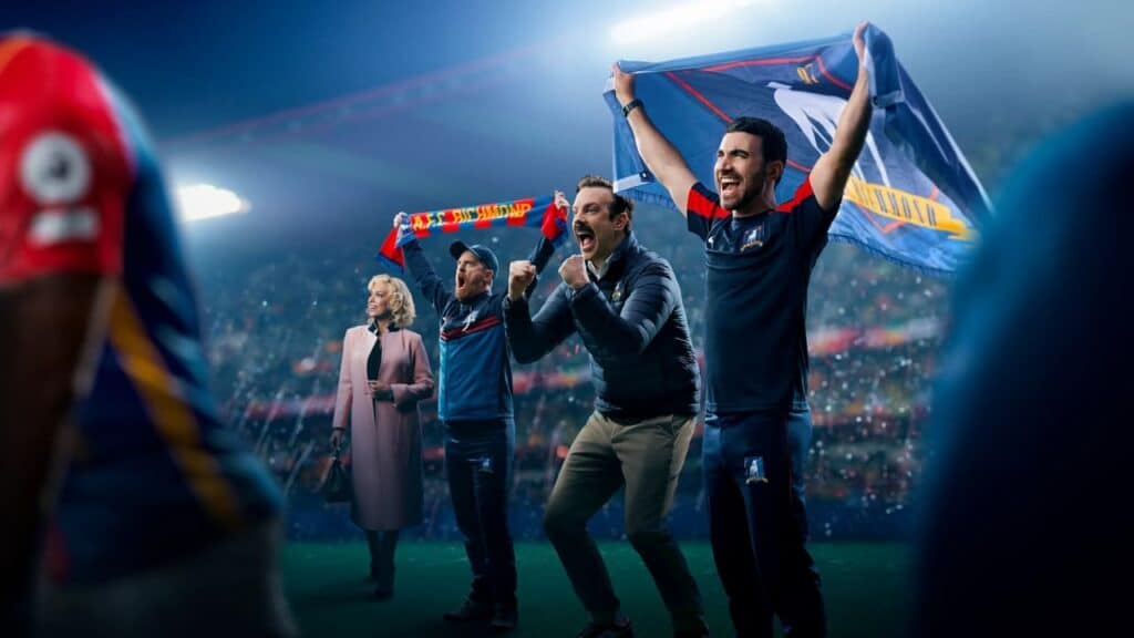 Several people celebrate enthusiastically on a soccer field at night, cheering and holding team scarves in a packed stadium.