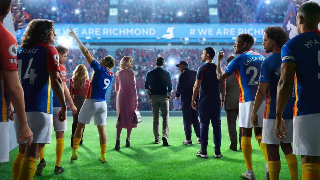 A soccer team and coaching staff walk onto a brightly lit field from a tunnel as fans cheer in a crowded stadium.