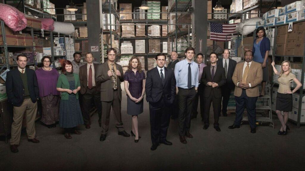 Cast of The Office standing in a Dunder Mifflin warehouse setting, reflecting the show’s grounded workplace atmosphere and mockumentary style.