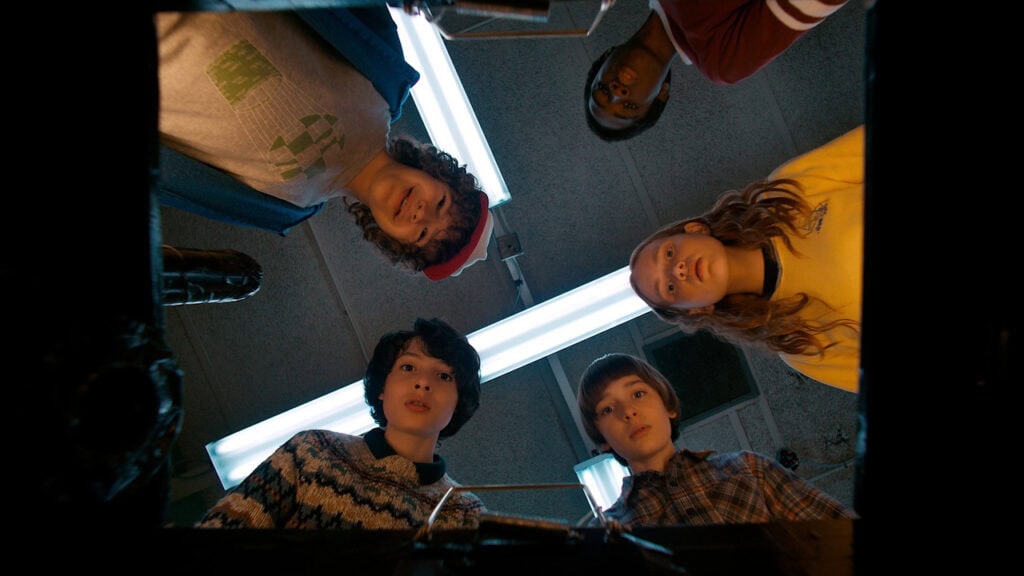 A group of kids from Stranger Things look down into an open space with concerned expressions as bright lights shine above them.