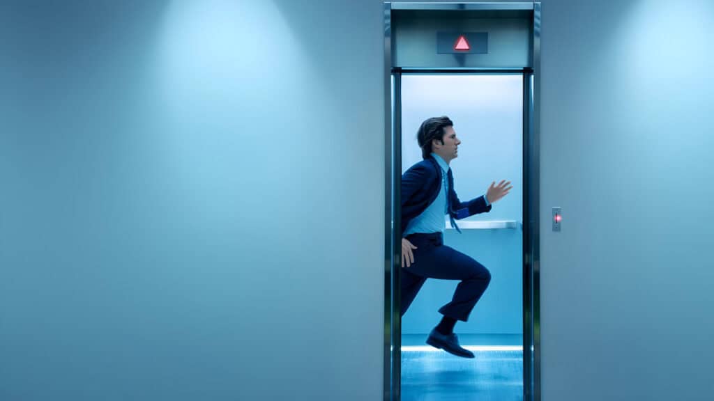 A man in a suit running out of an elevator into a sterile blue hallway representing urgency and control inside the Severance workplace