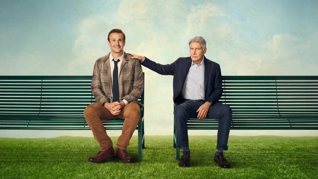 Two men sit on a green park bench on a grassy field, one resting a supportive hand on the other’s shoulder beneath a soft, cloudy sky.