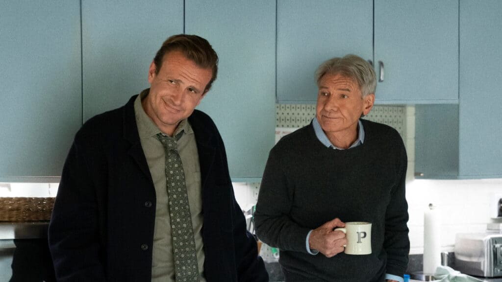 Two men lean against a kitchen counter in a light blue kitchen, one holding a mug while they exchange an amused, knowing look.
