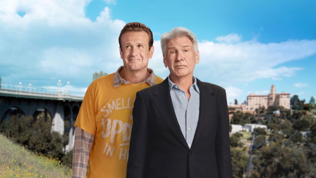 Two men stand side by side outdoors near a bridge and hillside neighborhood, one smiling casually while the other looks serious against a bright blue sky.