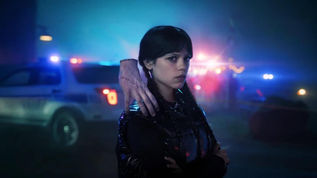 Wednesday Addams stands with a stitched hand on her shoulder as flashing police lights glow behind her during a nighttime investigation.