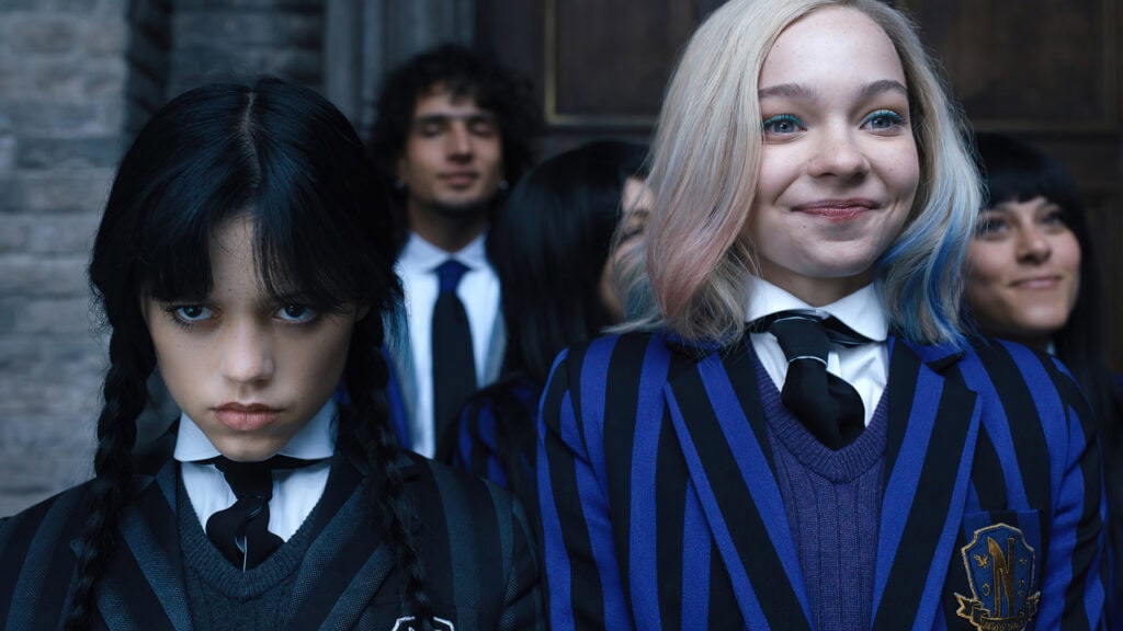 Wednesday Addams stands serious beside her cheerful roommate Enid as they wear their Nevermore Academy school uniforms outside the academy.