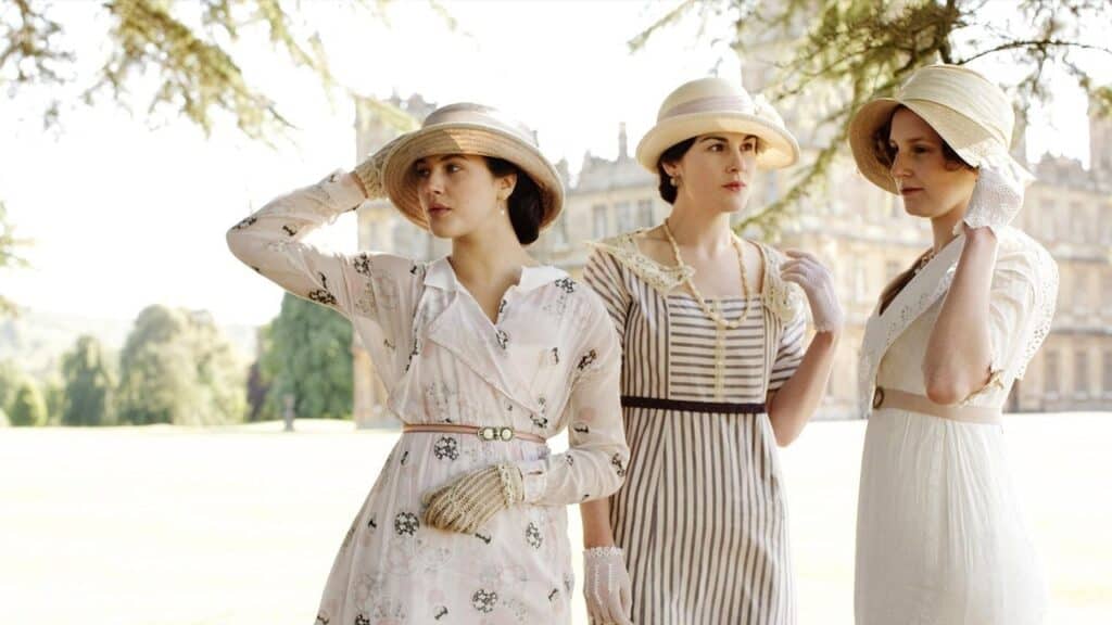 Three Downton Abbey characters wearing early 20th century Edwardian dresses and hats standing on the estate grounds, reflecting the show’s historical realism.
