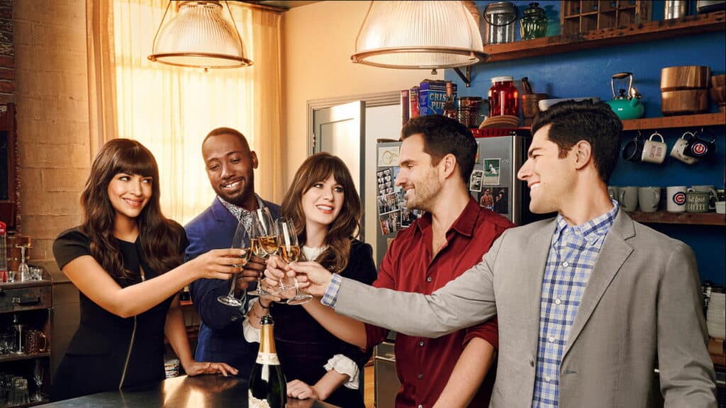 Five friends dressed up raise champagne glasses for a toast in a warm loft kitchen scene from New Girl.