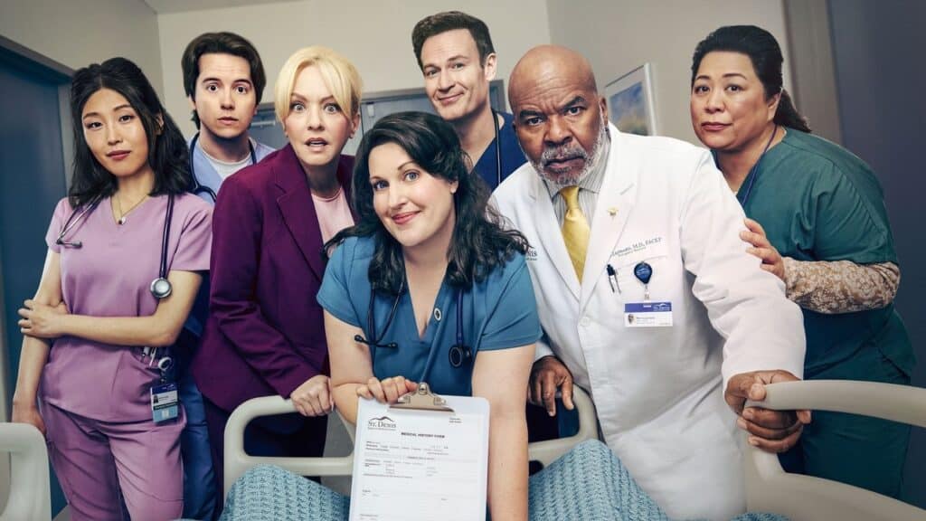 A wide-angle, full-cast photograph for the series St. Denis Medical. Seven diverse cast members, in a range of blue, pink, and green scrubs and professional wear, group closely around the foot of a hospital bed, looking down toward the perspective of a hypothetical patient. In the center, Allison Tolman's character holds up a medical history form on a clipboard. They have diverse expressions of professional curiosity, concern, and quirkiness. The setting is a brightly lit hospital room with blue doors.