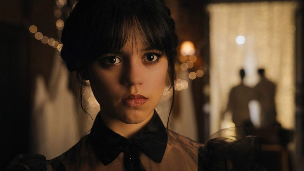 Close-up still of Wednesday Addams in a dark formal outfit, standing indoors with warm lights and blurred figures in the background.