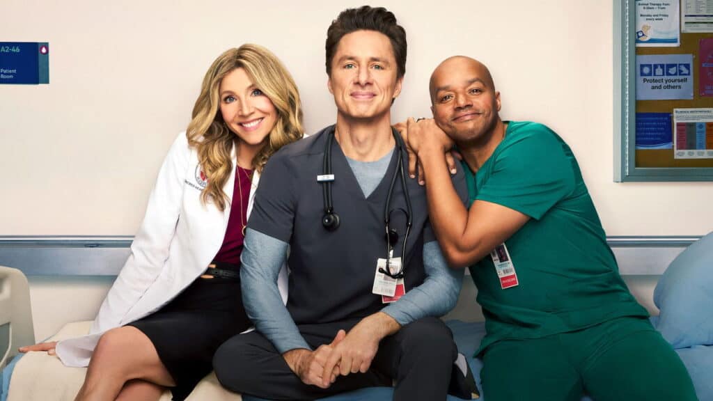 A horizontal landscape picture of Sarah Chalke, Zach Braff, and Donald Faison, wearing medical scrubs and a white coat, sitting side by side on a hospital bed and smiling at the camera. J.D. and Turk are holding hands and there is a hospital wall behind them with patient room signs and a bulletin board.