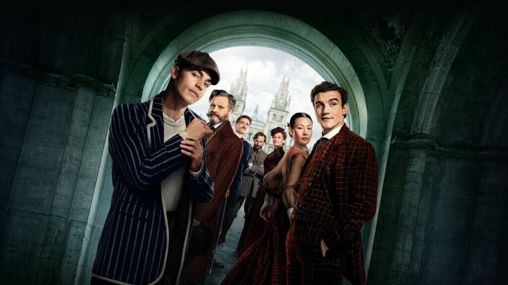 A group photo of the Young Sherlock cast members standing in front of an ancient stone archway leading to Oxford-style buildings. The cast features several recognizable actors, including Hero Fiennes Tiffin as Young Sherlock and Colin Firth, all dressed in 19th-century attire.