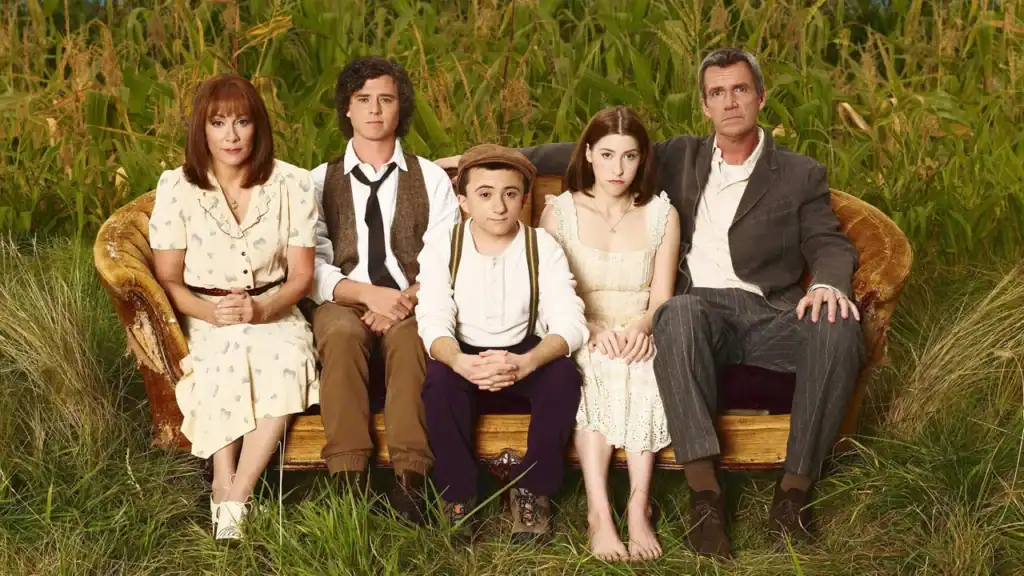 An official promotional poster for ABC's The Middle. In a deliberate twist on a normal cast shot, the five members of the Heck family (Frankie, Brick, Sue, Axl, and Mike) pose on an old, dilapidated, frayed brown couch sitting in the middle of a sparse field of tall corn stalks and wild grass. They are wearing dated, dusty, 1930s-style blue-collar and farming clothes. The family looks directly at the camera with serious, unglamorous, and almost judgmental expressions, capturing the show’s authentic, struggling small-town vibe.
