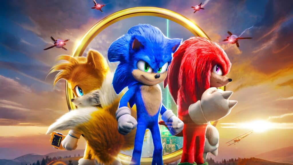 Wide cinematic shot of Tails, Sonic, and Knuckles standing together in front of a large golden ring with a sunset and flying drones in the background.